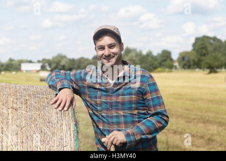 Agricoltore appoggiata contro la balla di fieno guardando sorridente della fotocamera Foto Stock