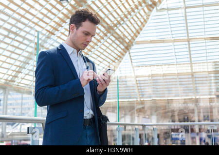 Imprenditore iscritto un messaggio sullo smartphone, stazione ferroviaria in attesa lounge in background Foto Stock