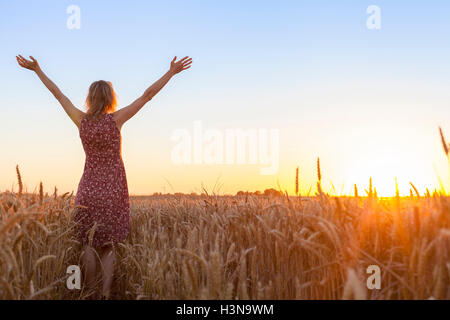 Positivo felice donna piena di vitalità alzando le mani e di fronte al sole in un campo di grano di sunrise Foto Stock