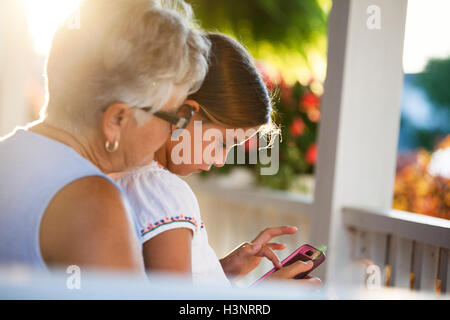 Ragazza che gioca il gioco dello smartphone seduto su alla nonna giro in veranda al tramonto Foto Stock