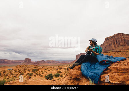 Giovane uomo seduto su rock suonare la chitarra acustica, Monument Valley, Arizona, Stati Uniti d'America Foto Stock