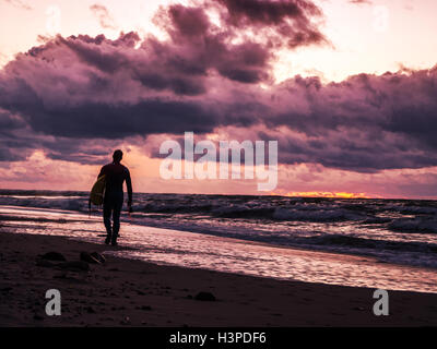 Surfer passeggiate lungo la spiaggia al tramonto Foto Stock