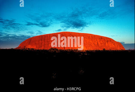 Alba a Uluru provoca la terra-cotta-superficie colorata di questo mondo-famoso monolito di pietra arenaria a comparire in rosso fiammante luce in mezzo la outback scrublands del Territorio del Nord in Australia. Noto anche come Ayers Rock, la massiccia landmark sorge a circa 350 metri (1148 piedi) ) al di sopra del suolo e coperchi 8,6 chilometri quadrati (3.32 miglia quadrate) in Uluru-Kata Tjuta National Park. Si tratta di un luogo sacro per la zona indigene tribù aborigena, la Anangu, che ha chiamato le antiche rocce Uluru, il significato di "Terra Madre". Foto Stock