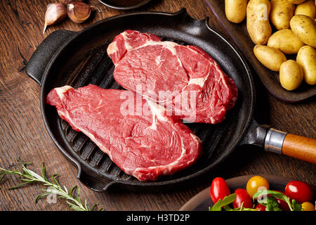 Materie bistecca in padella per grigliare, patate, spezie e pomodori sul tavolo di legno Foto Stock