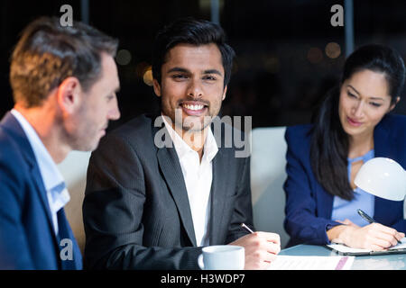 Imprenditore sorridente alla telecamera mentre i colleghi guardando il documento Foto Stock