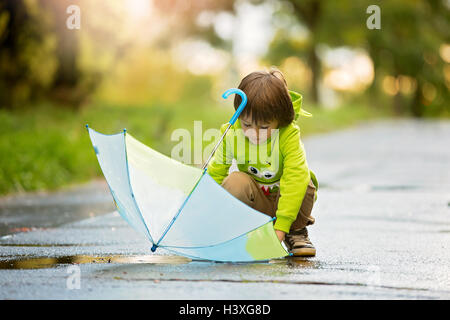 Adorable little boy con ombrello in un parco in un giorno di pioggia, giocando e saltando, sorridente, tempo di autunno Foto Stock