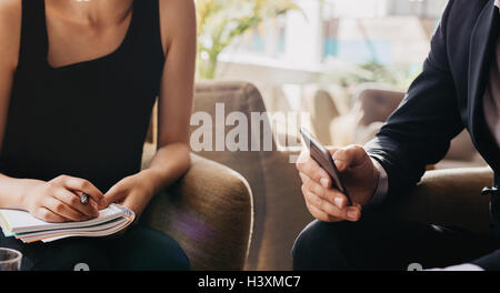 Immagine ritagliata di imprenditore tenendo il telefono cellulare e la donna con il blocco note. Gli imprenditori seduti nella hall. Foto Stock