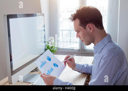 Giovane imprenditore lavora con i report finanziari in un ufficio moderno con tavolo in legno e computer con schermo vuoto per copyspace Foto Stock