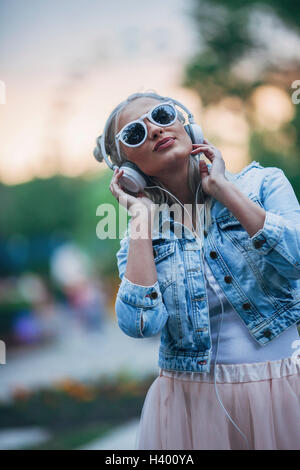 Moda giovane donna che indossa gli occhiali da sole mentre si ascolta la musica attraverso le cuffie Foto Stock