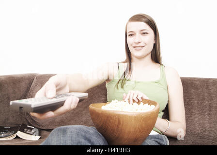 Camere salotto, divano, donna, giovane, sedersi, tenere ciotola, telecomando, gesto, zap ritratto a ginocchiera 22 anni, i giovani e gli adolescenti a pelo lungo, dai capelli scuri, tempo libero e week-end, di essere pigro, attività per il tempo libero, televisione, media, intrattenimento, scorza di legno, popcorn, lettino, all'interno, a casa Foto Stock