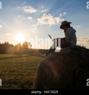 Mari uomo seduto sulla balla di fieno al tramonto con laptop Foto Stock