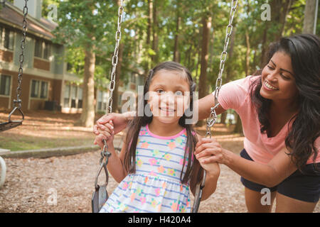 Madre ispanica spingendo la figlia nel parco giochi swing Foto Stock