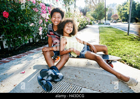 Razza mista del fratello e sorella seduta su skateboard sul marciapiede Foto Stock