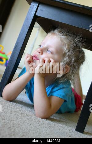 Bambino o bambina che si nasconde sotto un piccolo tavolo guardando fuori della finestra Foto Stock