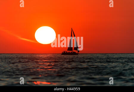 Barca a vela al tramonto si profila una barca con equipaggio a bordo navigando lungo l'acqua oceanica con un bianco caldo sole che tramonta il sole rosso Foto Stock