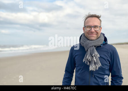 Sorridente uomo di mezza età con gli occhiali e una lana lavorata a maglia Sciarpa in piedi su una deserta spiaggia di autunno in un giorno nuvoloso con cop Foto Stock