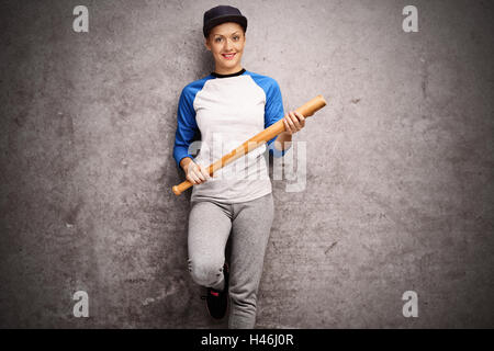 Atleta femminile appoggiata contro un arrugginito muro grigio e in possesso di una mazza da baseball Foto Stock