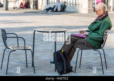 Un anziano uomo seduto da lui stesso facendo un cruciverba in cortile della Somerset House, Londra. Foto Stock