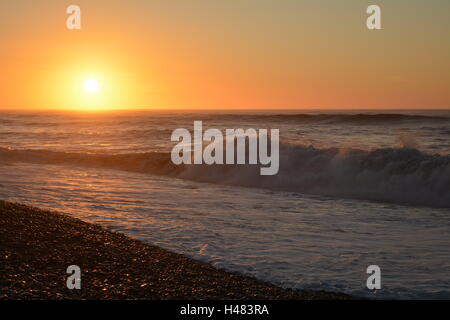 Le onde si infrangono sulla riva con una luminosa arancione tramonto Foto Stock