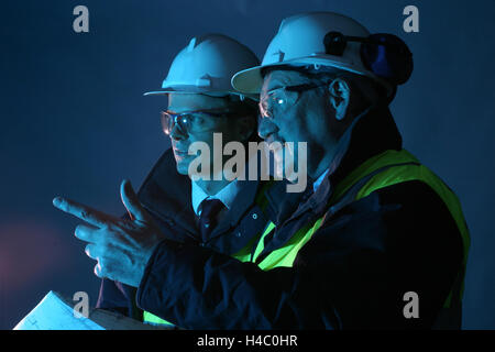 2 tecnici senior (una giovane, uno più vecchio) discutere il lavoro durante il puntamento. Indossare copricapi rigidi, ad alta viz canottiere e occhiali di sicurezza Foto Stock