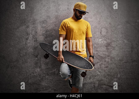 Pattinatore tenendo un longboard e appoggiata contro un arrugginito muro grigio Foto Stock