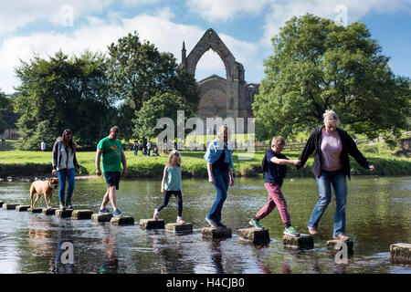 I membri del pubblico attraversare il fiume Wharfe insieme con il loro cane, mentre vi godete il sole, a Bolton Abbey, in Wharfedale, Foto Stock