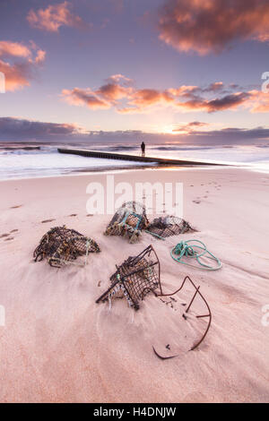Fotografo solitario sul pontile a Blyth beach, con aragosta padelle e pentole di granchio mezzo sepolto nella sabbia al sorgere del sole in inverno Foto Stock