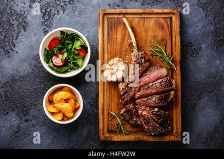 Fette di bistecca alla griglia su ossa bovine nervatura con spicchi di patate e insalata con pomodori e rucola su sfondo scuro Foto Stock