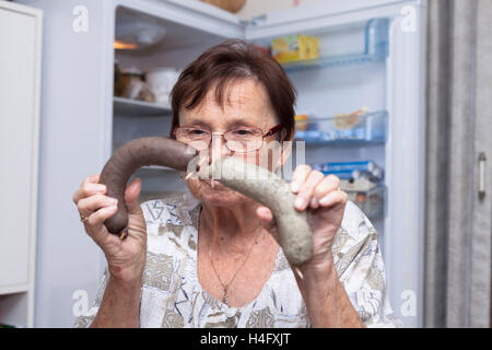 Senior donna holding salsicce di fegato di maiale mentre in piedi di fronte ad aprire il frigorifero in cucina. Foto Stock