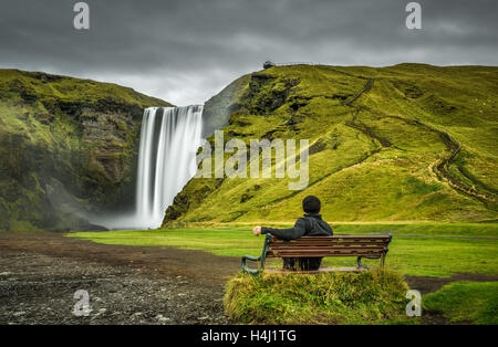 Escursionista si siede su una panchina e guarda alla famosa cascata Skogafoss nel sud dell'Islanda Foto Stock