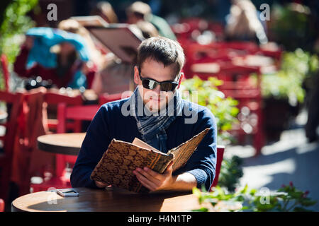 Giovane uomo bello indossare occhiali da sole leggendo il giornale del mattino in una street cafe guardando lateralmente. Pausa caffè. Foto Stock
