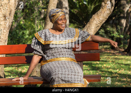 Ritratto di una donna africana indossando il tradizionale abito seshweshwe seduto su una panca di legno in un giardino rilassante Foto Stock
