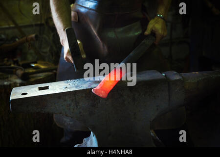 Un fabbro colpisce una lunghezza di rosso metallo caldo sull'incudine con un martello in un workshop. Foto Stock