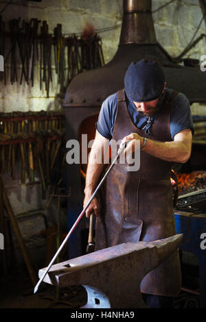 Un maniscalco in un grembiule in pelle trattiene una lunghezza sottile di metallo contro l'incudine in un workshop. Foto Stock