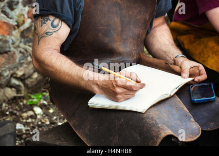 Due persone, un uomo e una donna fabbri indossando grembiuli di cuoio la scrittura in un notebook sat in un giardino. Foto Stock