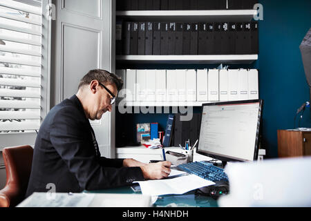 Un uomo seduto alla scrivania in ufficio, scritto su un pezzo di carta. File ordinate di file su ripiani e carte sulla scrivania. Foto Stock