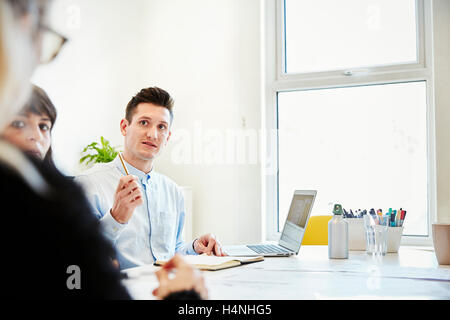 Un uomo che utilizza un computer portatile seduti ad un tavolo a parlare con due colleghi in ufficio. Foto Stock