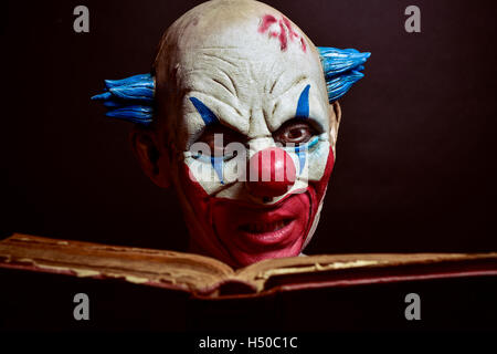 Primo piano di una paura male clown la lettura di un vecchio libro, contro uno sfondo scuro Foto Stock