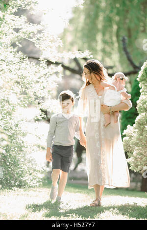 Ritratto di una madre sorridente, bambino e bambina con una ghirlanda di fiori sulla sua testa, camminare in un giardino. Foto Stock