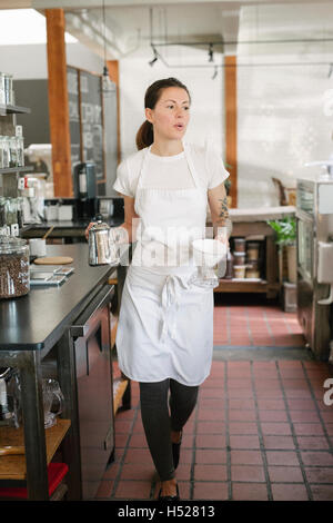 Donna che indossa il grembiule camminando lungo un corridoio che porta un POT del caffè e il filtro. Foto Stock