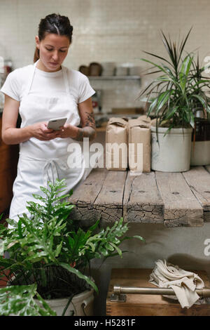 Donna che indossa un grembiule bianco in una cucina, utilizzando un telefono cellulare. Foto Stock