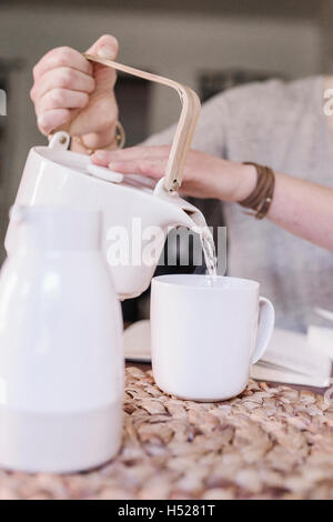 Donna seduta al tavolo nel suo appartamento, versando acqua calda in una tazza, mattina di routine. Foto Stock