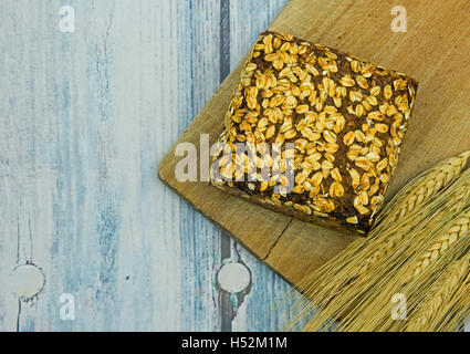 Piccola pagnotta di pane con semi e fiocchi di avena, alcune spighe di grano nella scheda da cucina in legno e bancone blu in un vintage Foto Stock