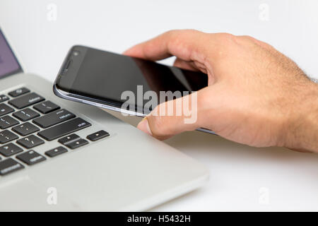 Uomo con uno smartphone nella sua mano destra con il computer portatile in backgroud sia spento. studio shot su sfondo bianco Foto Stock