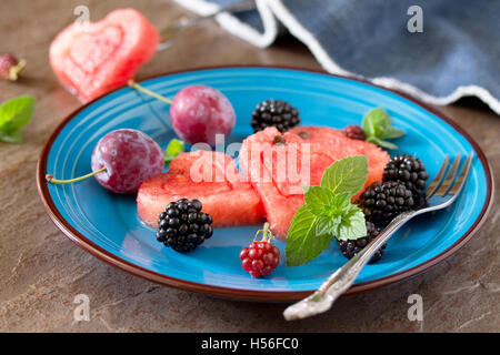 Fetta di cocomero cuore e frutti di bosco (blackberry, prugna) sulla pietra marrone dello sfondo. Foto Stock