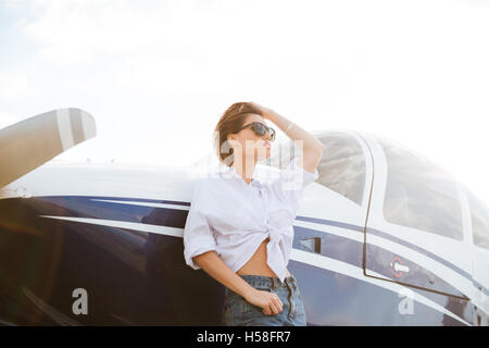 Bella giovane donna in occhiali da sole in piedi all'aperto nei pressi di piccoli aerei privati Foto Stock