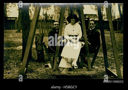 Una cartolina fotografica cattura due giovani uomini e una donna su un'altalena di legno, fornendo un'istantanea delle attività ricreative e dei costumi sociali durante il periodo in cui è stata scattata. Foto Stock