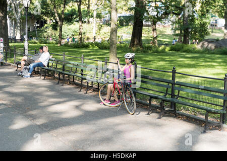 Giovane donna che indossa un top rosa e il ciclismo casco di sicurezza in appoggio su una panchina del Central Park di New York City tra gite in bicicletta Foto Stock