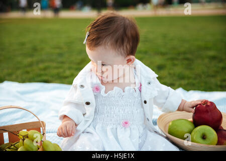 Incantevole piccola ragazza seduta e scegliendo la frutta in picnic Foto Stock