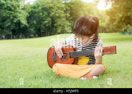 Asian bimbi piccoli ragazza seduta su erba e giocare ukulele nel parco. Kids Giochi divertenti ukulele Foto Stock